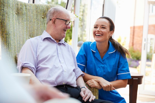 A young female caregiver attends to a senior man sitting in a chair.