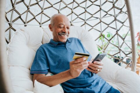 A senior man sits on a wicker chair and shops on a smartphone.